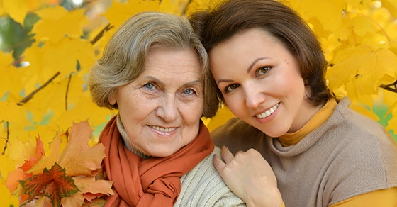 An adult woman and her elderly mother pose for a photo amidst yellow leaves.