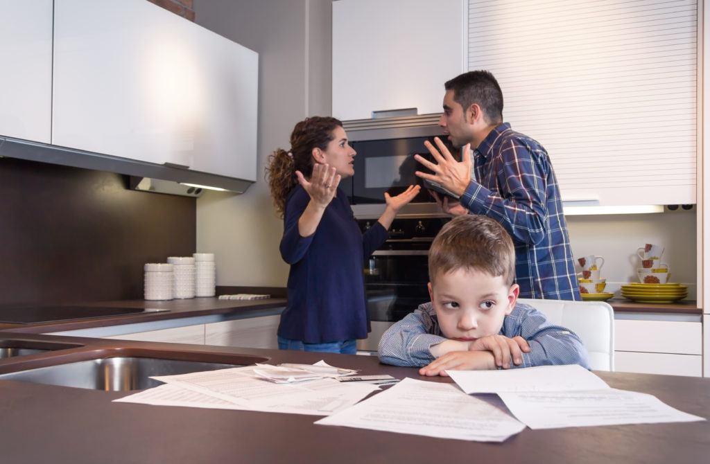 A mother and father argue while while an upset son sits at a kitchen counter.