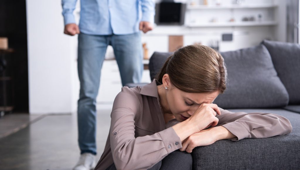 A woman on a couch cries while a man stands in the background with their fists clenched.
